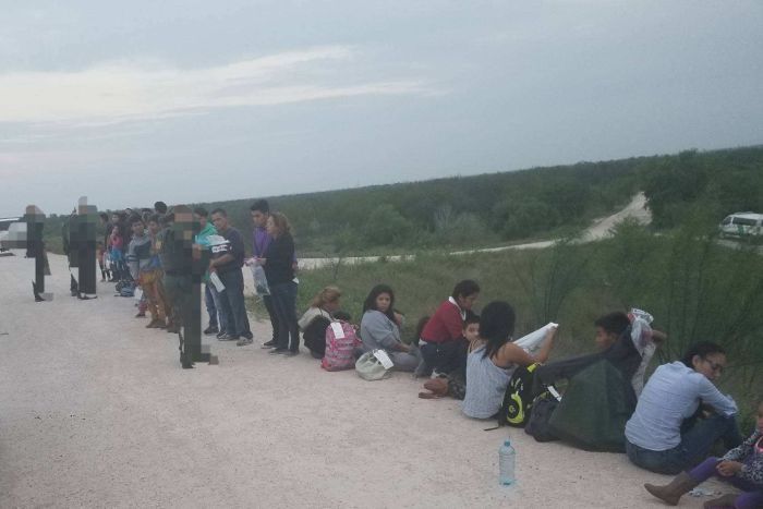 A line of people sit on the side of the road, some stand talking to officers in uniform.